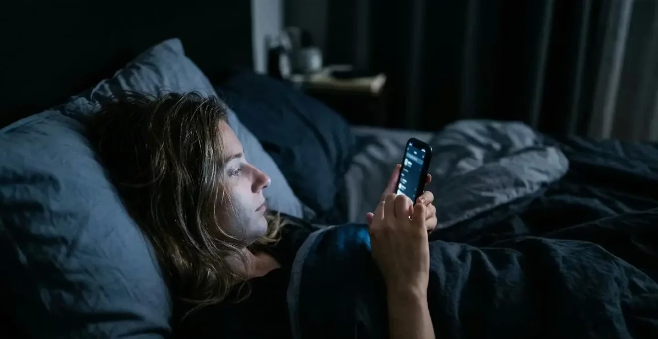 Person scrolling smartphone in dark bedroom with blue screen glow affecting melatonin production